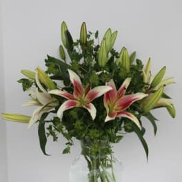 Tall arrangement of pink and white lilies with many buds in a clear glass vase