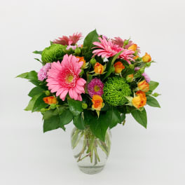 Bright bouquet of pink gerbera daisies, orange roses, and green mums in a clear glass vase