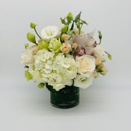 Round arrangement of white hydrangeas, cream roses, and orchids in a glass vase