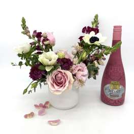 Mixed pink and white flower arrangement in white vase beside pink rosé bottle and scattered petals
