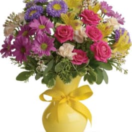 Mixed bouquet of pink, purple, and yellow flowers in a yellow vase with a ribbon