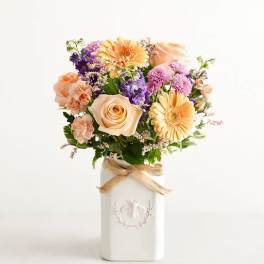 Peach and purple mixed flower arrangement in a white ceramic vase with a burlap bow