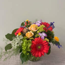 Mixed bouquet with red gerbera daisies, peach roses, and purple flowers in a glass vase