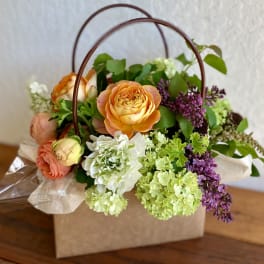 Peach roses, green hydrangeas, and purple flowers in a kraft box with curved handles
