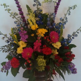 Tall mixed flower arrangement in a dark pedestal vase with bright pink, yellow, blue, and purple blooms