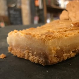 A square dessert bar with a crumbly golden crust on a dark surface