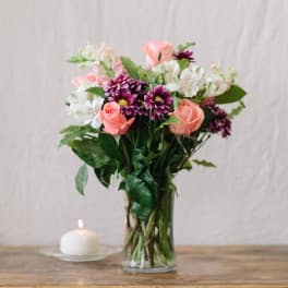 Pink roses and purple daisies in a clear glass vase beside a lit candle.