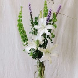Tall white lily arrangement with green and purple spikes in a glass vase