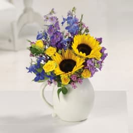 Bouquet of sunflowers, yellow roses, and purple flowers in a white pitcher vase