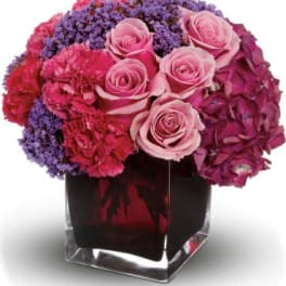 Compact arrangement of pink roses, carnations and purple blooms in a clear square glass vase
