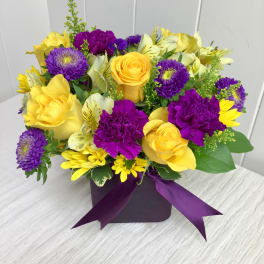 Low cube arrangement of yellow roses and daisies with purple carnations and asters in a purple vase with ribbon