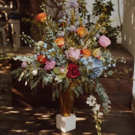 Tall floral arrangement in a gold vase with pink, orange, blue, and white blooms