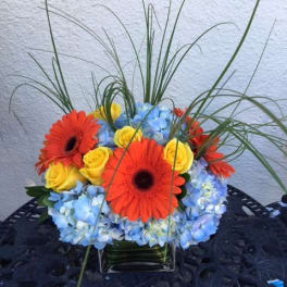 Bright bouquet of orange gerbera daisies, yellow roses, and blue hydrangeas in a glass vase