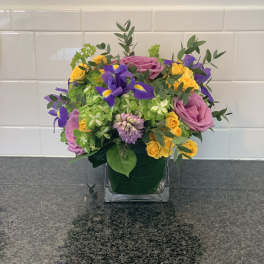 Mixed bouquet of pink roses, yellow spray roses, and purple irises in a square glass vase
