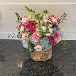 Mixed bouquet of pink roses, blue hydrangeas, and white blooms in a wrapped container