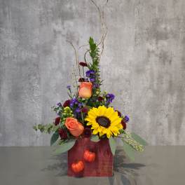 Tall floral arrangement with sunflowers, orange roses, and purple blooms in a red box