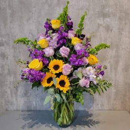 Tall bouquet of yellow sunflowers, lavender roses, and purple flowers in a glass vase
