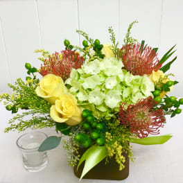 Low arrangement of green hydrangea, yellow roses, and red pincushion protea in a square vase with a small votive candle.