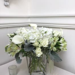 White floral arrangement in a clear square vase with calla lilies and roses
