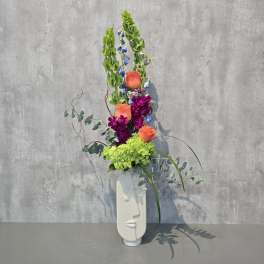 Tall floral arrangement with orange roses, purple blooms, and green hydrangea in a white vase