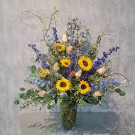 Tall bouquet of sunflowers, blue flowers, and peach roses in a glass vase