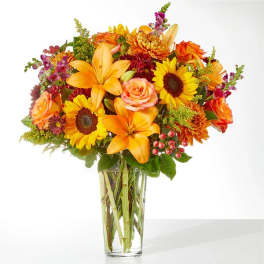 Mixed bouquet of orange lilies, sunflowers, and roses in a clear glass vase