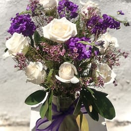 White roses and purple flowers arranged in a glass vase with ribbon