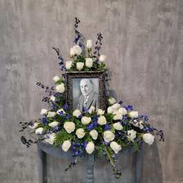 White roses and blue flowers arranged around a framed portrait on a table