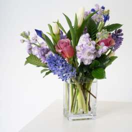 Mixed bouquet of pink roses, purple stock, blue blooms, and white tulips in a clear glass vase