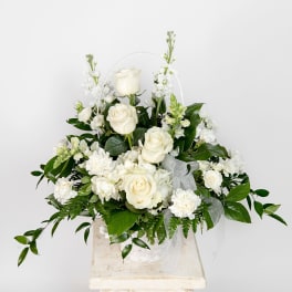 White floral basket with roses and carnations arranged with lush foliage and a sheer ribbon bow.