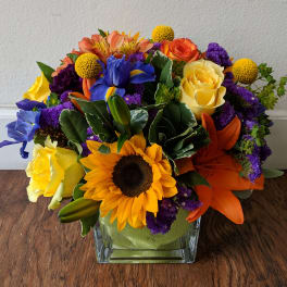 Colorful bouquet with sunflowers, roses, lilies, and purple filler in a square glass vase