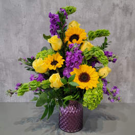 Purple vase holding yellow roses, sunflowers, green hydrangeas, and purple blooms