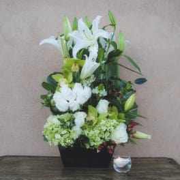 White lilies and green blooms in a black container beside a candle