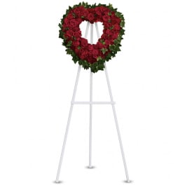 Heart-shaped wreath of red roses with a green foliage border on a tall white easel.