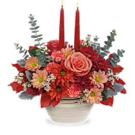 Red and peach floral arrangement in a white bowl with two red candles