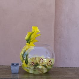 Yellow orchids arranged in a round glass bowl with a small square candle holder beside it