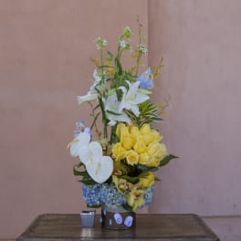 Tall floral arrangement with white lilies, yellow roses, and blue hydrangeas in a container