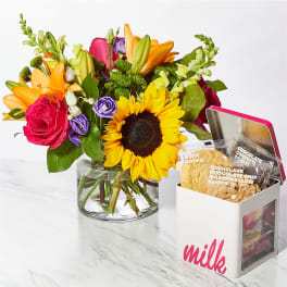 Bright mixed bouquet with sunflower, roses, and lilies beside a cookie tin on a marble surface