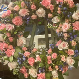 Standing wreath of pink roses and carnations with blue accent flowers on a green easel.