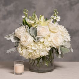 White roses and hydrangeas in a glass vase with a small candle beside it