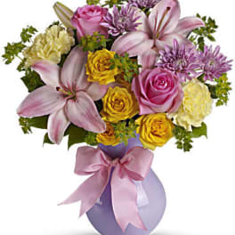 Pink and yellow mixed bouquet in a lavender vase with a pink ribbon