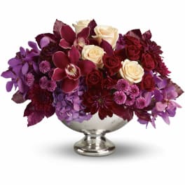 Low arrangement of cream roses, burgundy blooms, and purple flowers in a silver pedestal bowl