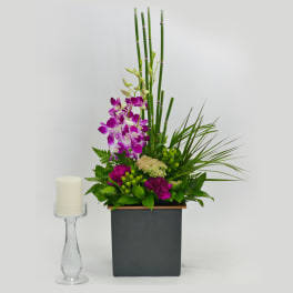 Tall orchid arrangement in a square black vase beside a white candle on a glass holder