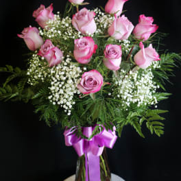 Pink roses in a glass vase with white filler flowers and a purple ribbon