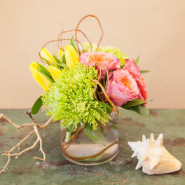 Pink and yellow flowers arranged in a clear glass vase with a seashell beside it