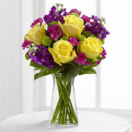 Yellow roses and purple flowers arranged in a clear glass vase