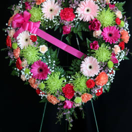 Heart-shaped floral wreath with a pink ribbon bow on a black stand