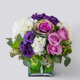 Compact arrangement of purple and lavender roses, lisianthus, and white hydrangeas in a square glass vase