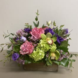 Low arrangement of pink roses, green hydrangeas, orchids and purple blooms in a rectangular wooden box