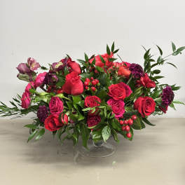 Low centerpiece of red and hot pink roses with accent blooms in a clear glass pedestal bowl.